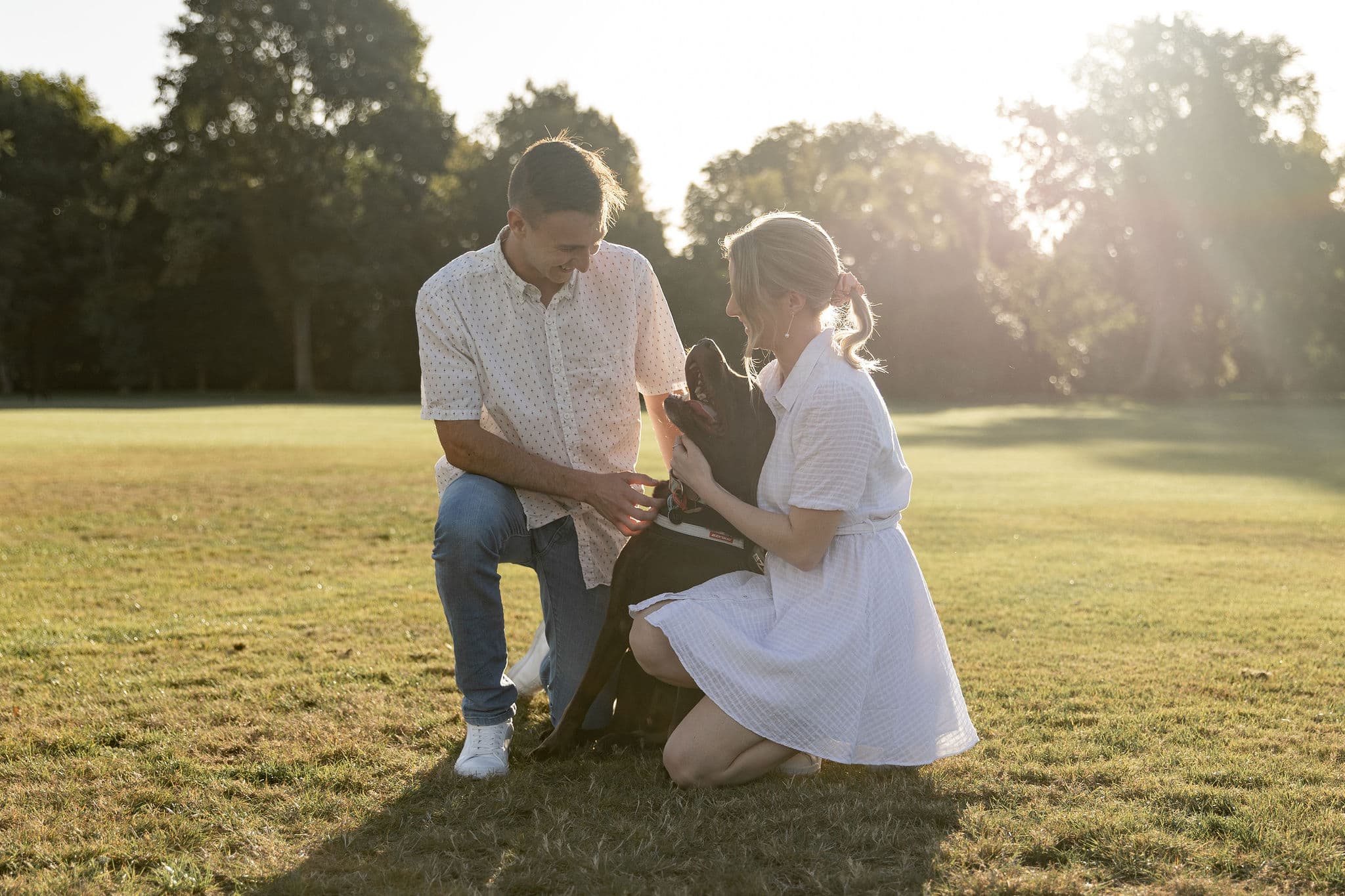 A couple kneels in a sunlit park, smiling at a black dog between them.