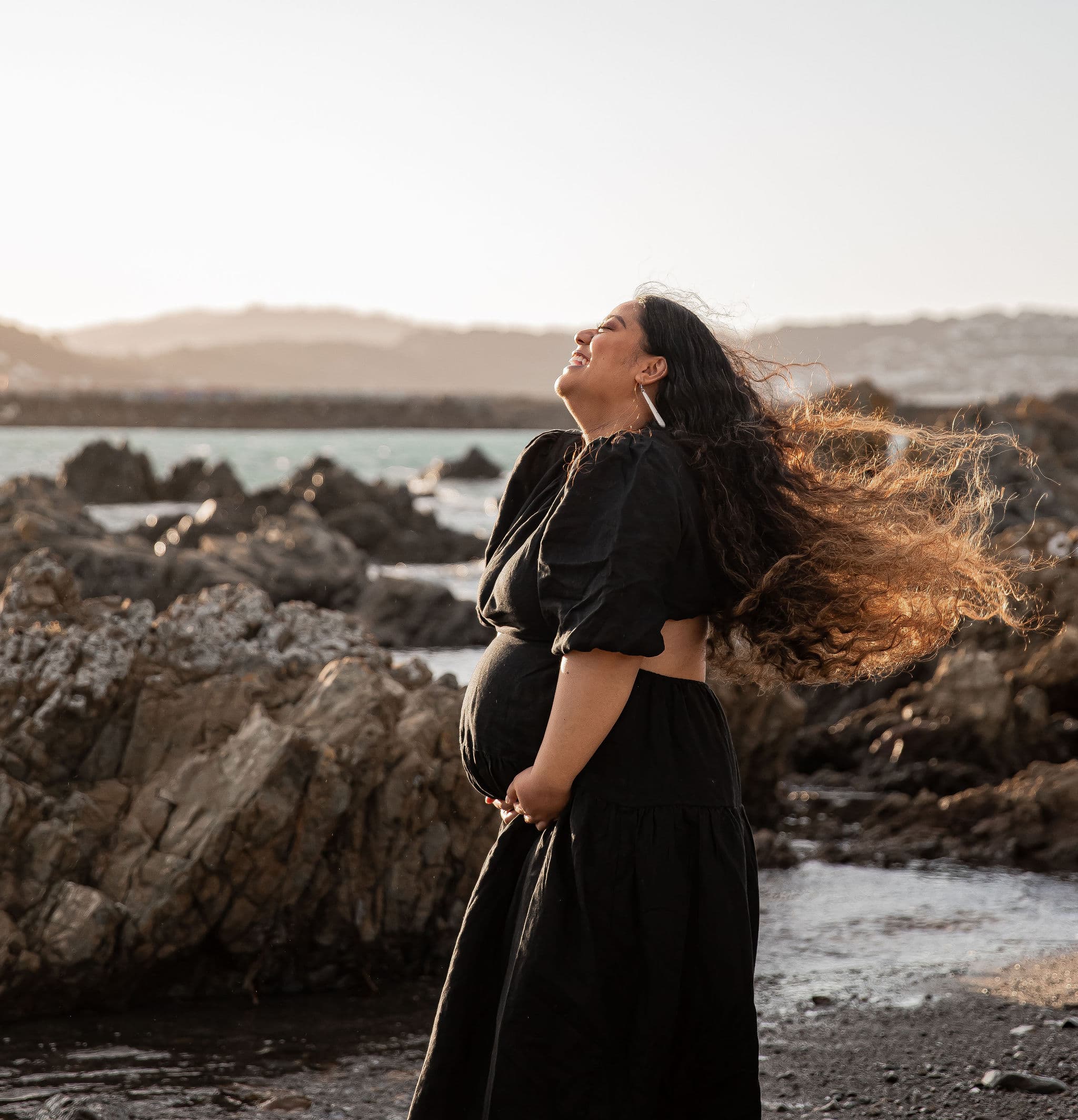 Smiling pregnant woman in a black dress stands by rocky shore with wind blowing her hair.