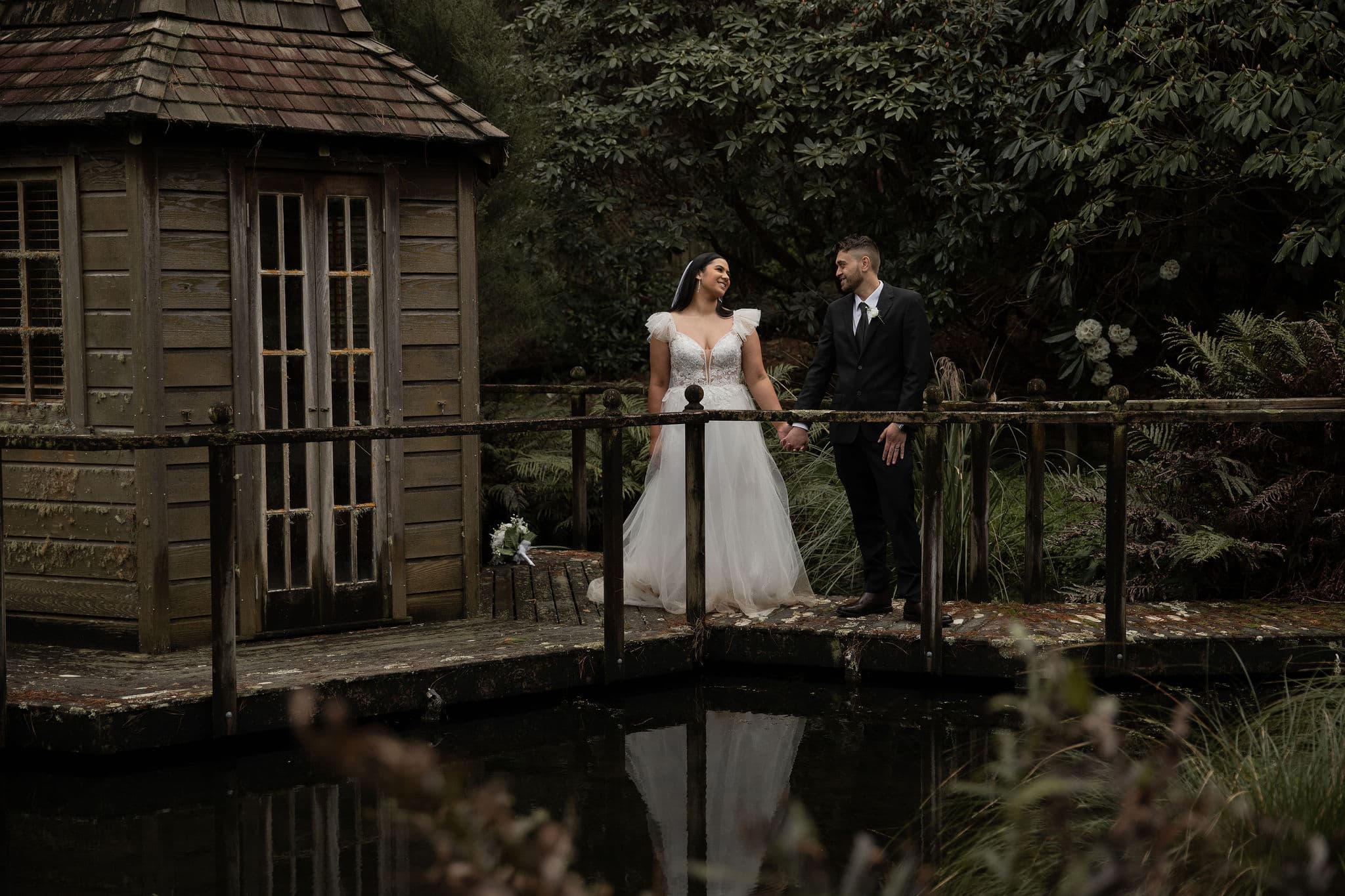 Bride and groom holding hands on a wooden bridge by a rustic shed and pond, surrounded by greenery.