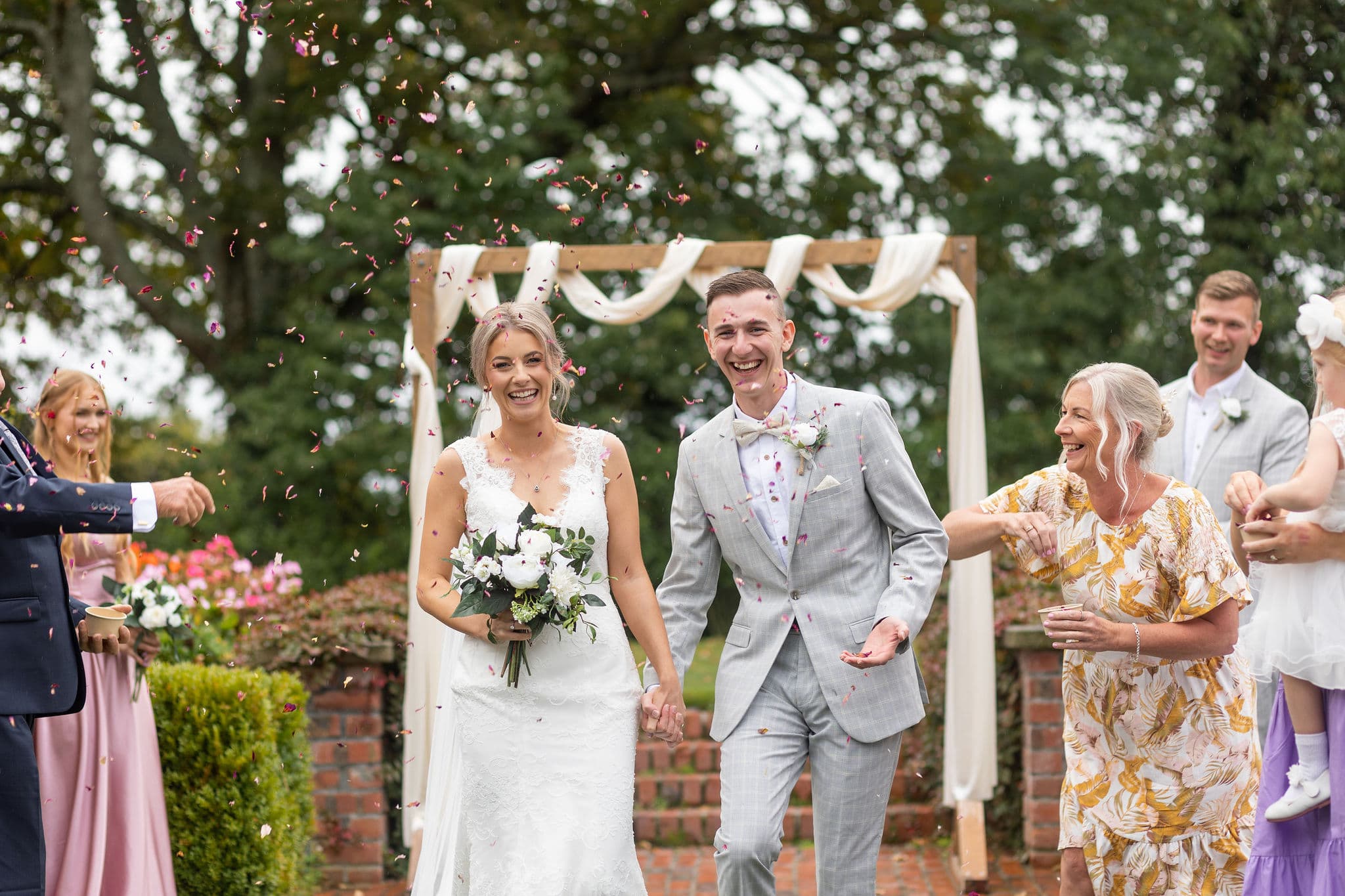 A bride and groom smile joyfully as confetti is thrown over them during an outdoor wedding ceremony, surrounded by happy guests. The bride holds a bouquet and the groom wears a light grey suit.