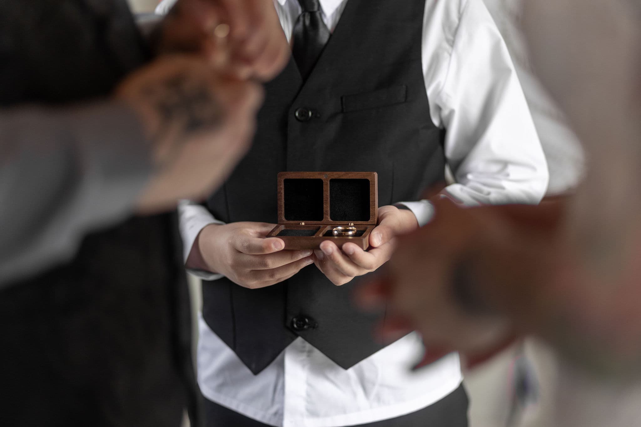 A person in a white shirt, black waistcoat, and tie holds an open wooden ring box with two rings inside, while others with blurred hands reach towards the box, suggesting a wedding ceremony.