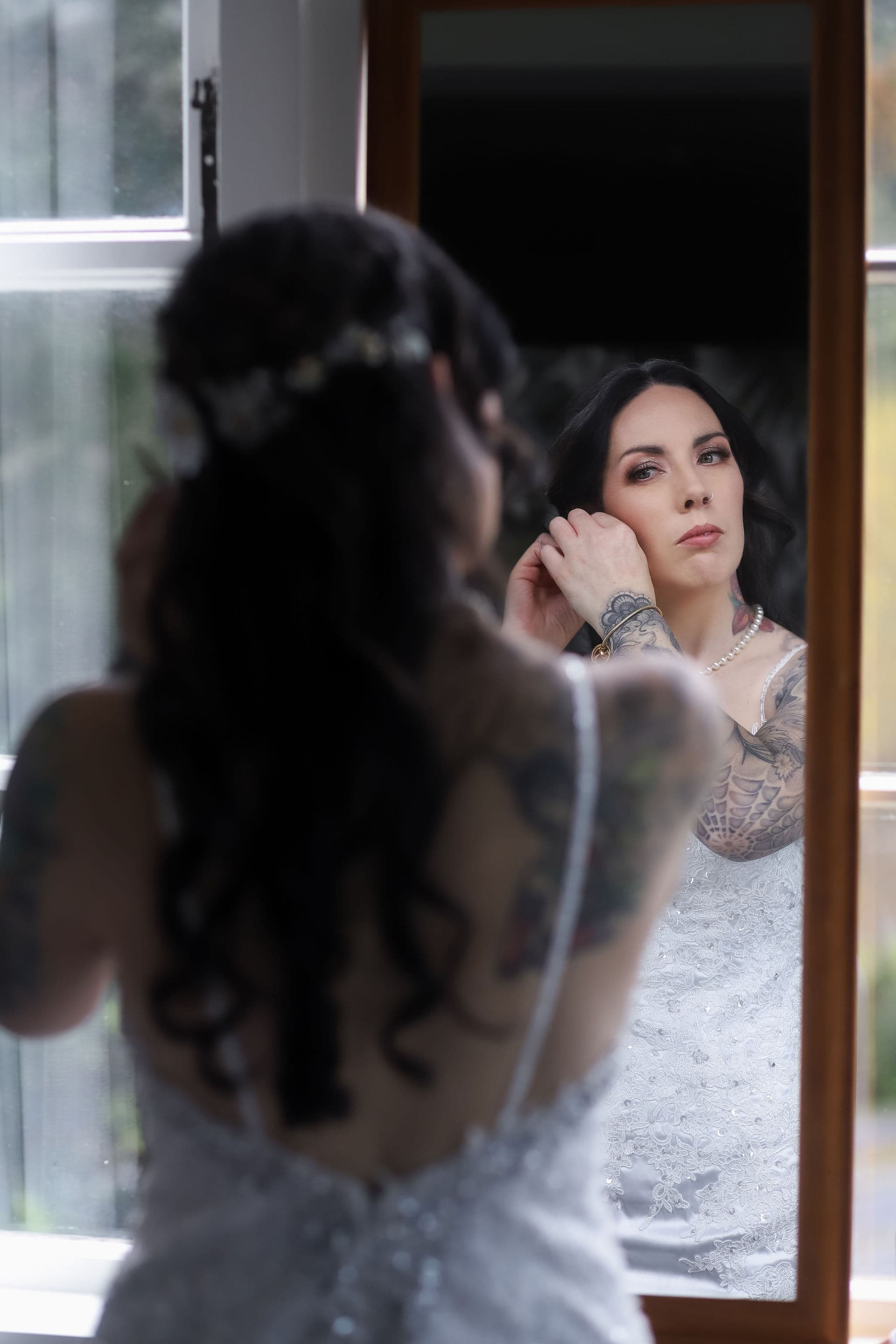 A woman with long dark hair, wearing a white dress, puts on an earring whilst looking at herself in a mirror. Her back faces the camera, and her reflection shows her focused expression. Tattoos are visible on her arms.