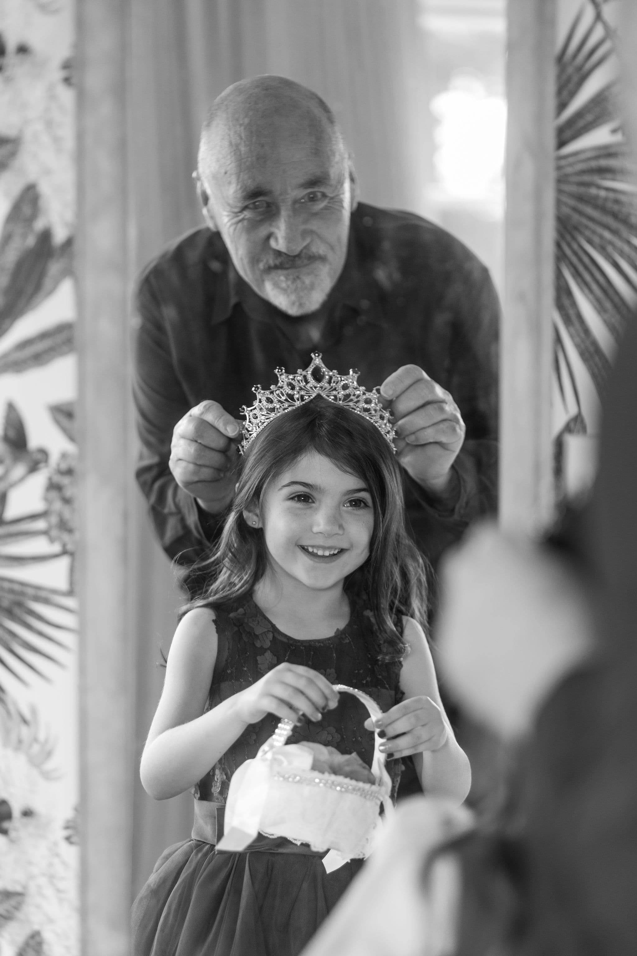 A smiling older man places a jewelled tiara on a young girl's head as she holds a small basket, both reflected in a mirror. The scene appears joyful and celebratory. Black and white photo.