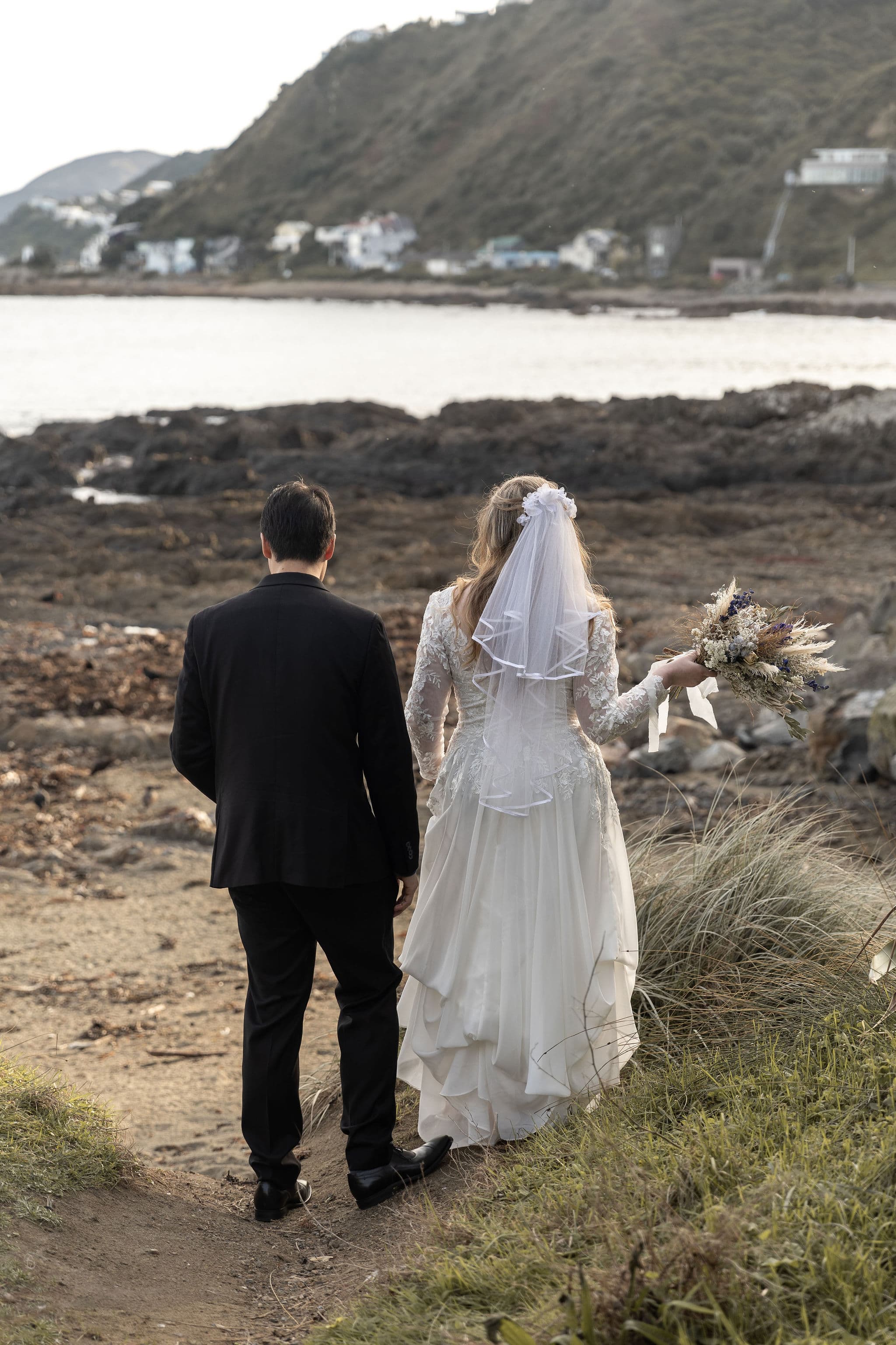 A bride and groom stand together, facing a rocky shoreline and distant hills.