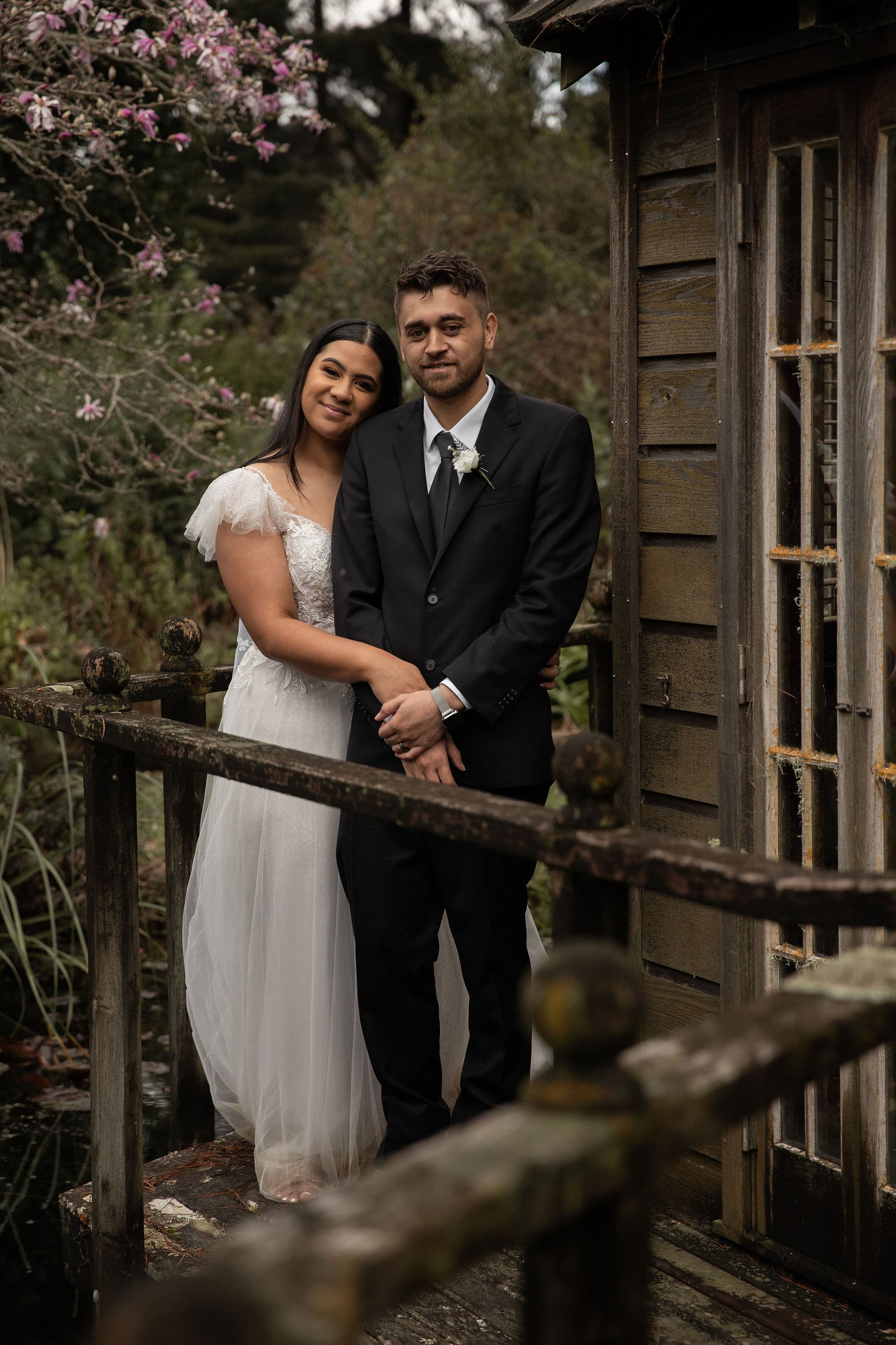 Bride and groom standing together on a rustic wooden bridge, smiling and cuddling.