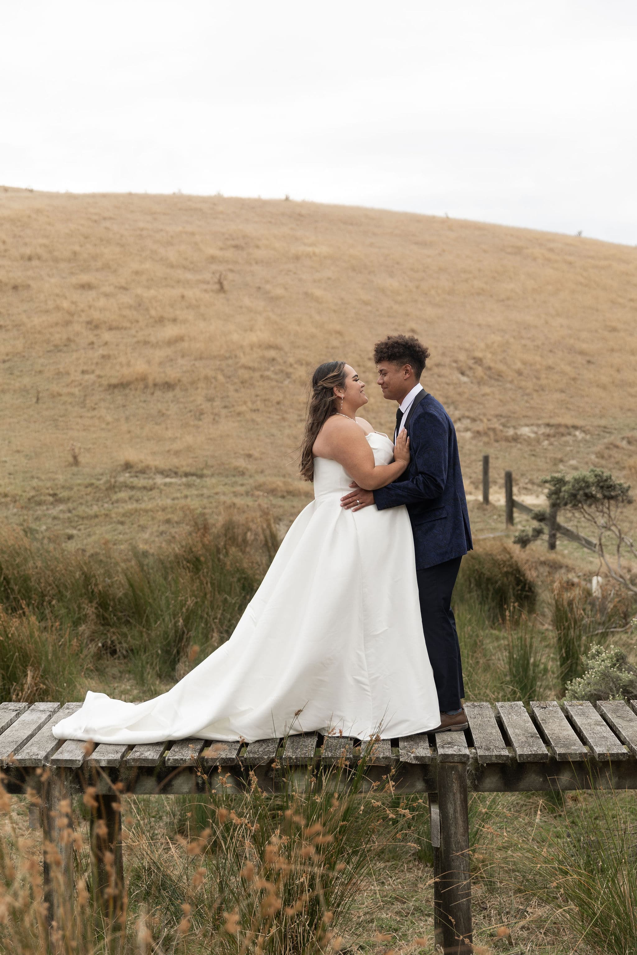 Bride and groom embrace on a wooden bridge in a grassy, hilly landscape.