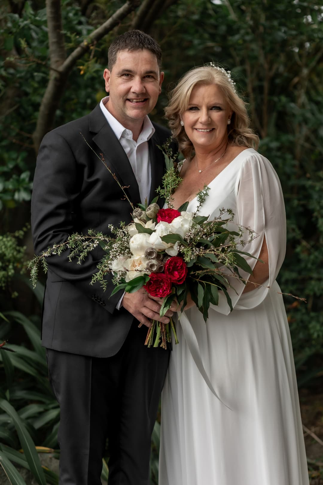A smiling couple pose outdoors; the woman wears a white dress and holds a bouquet of red and white flowers, whilst the man stands beside her in a black suit. Lush greenery surrounds them.