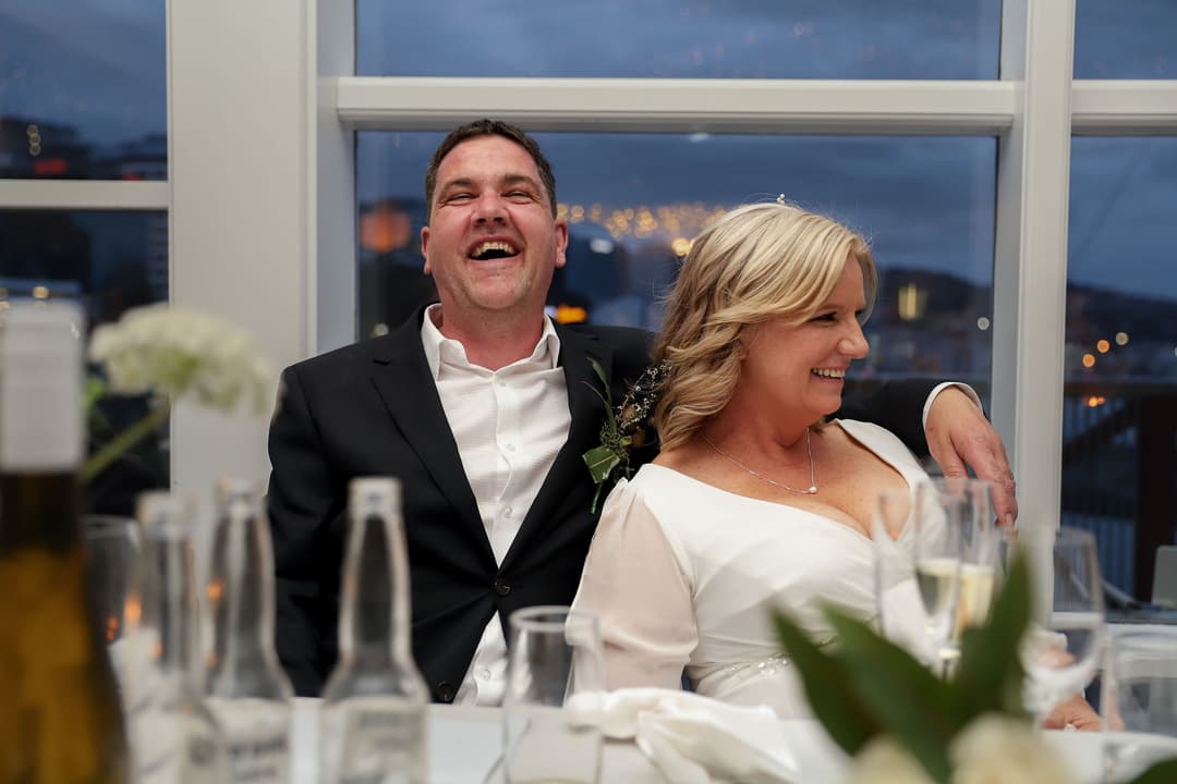 A smiling couple dressed formally sit closely together at a table, laughing and relaxed, with empty bottles and glasses in the foreground and city lights visible through the window behind them.