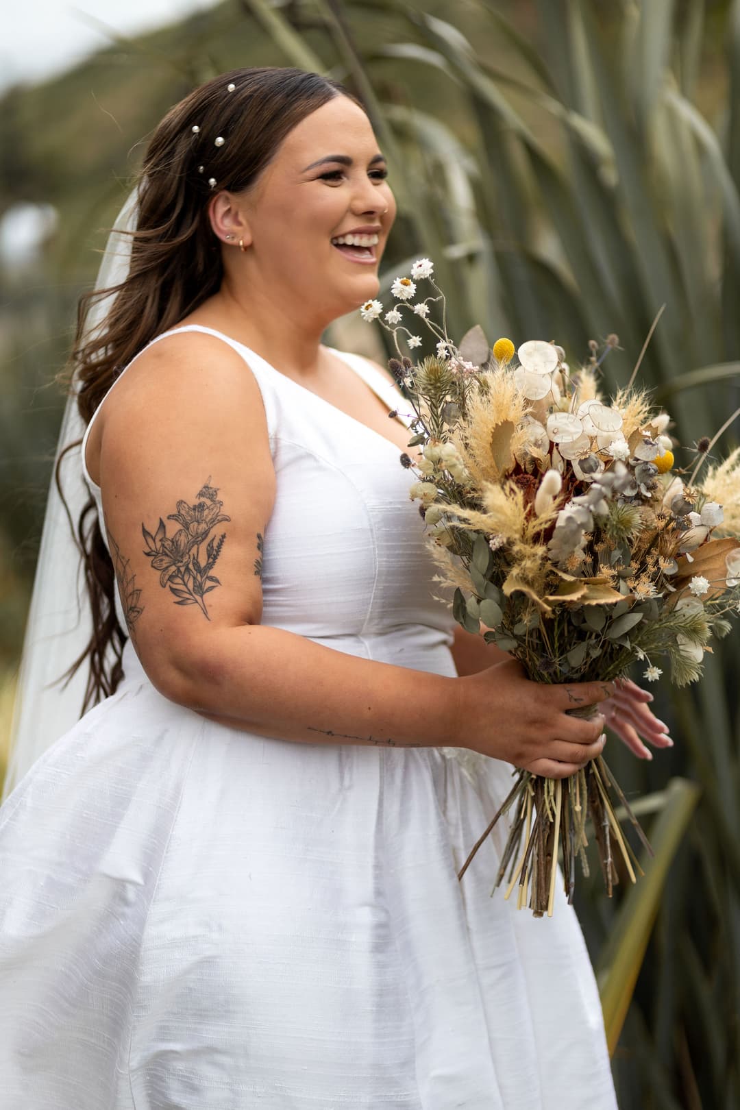 A smiling bride in a white dress holds a rustic bouquet of dried flowers; her long hair is loose with a veil and pearl accents, and she has a floral tattoo on her upper arm. Green foliage is blurred in the background.