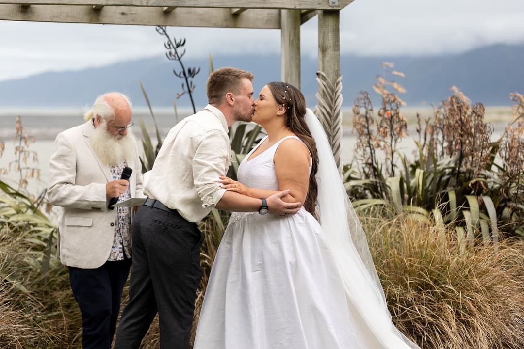 A bride and groom share a kiss during their outdoor wedding ceremony, standing under a wooden structure with greenery around them, while an officiant writes in a book nearby. Mountains and water are visible in the background.