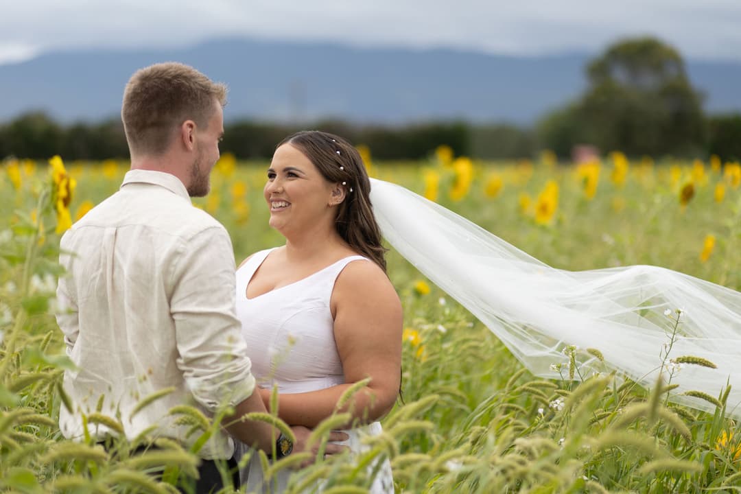 A bride and groom stand together in a field of sunflowers, smiling at each other. The bride wears a white dress and veil, while the groom wears a light shirt. Greenery and yellow flowers surround them under a cloudy sky.
