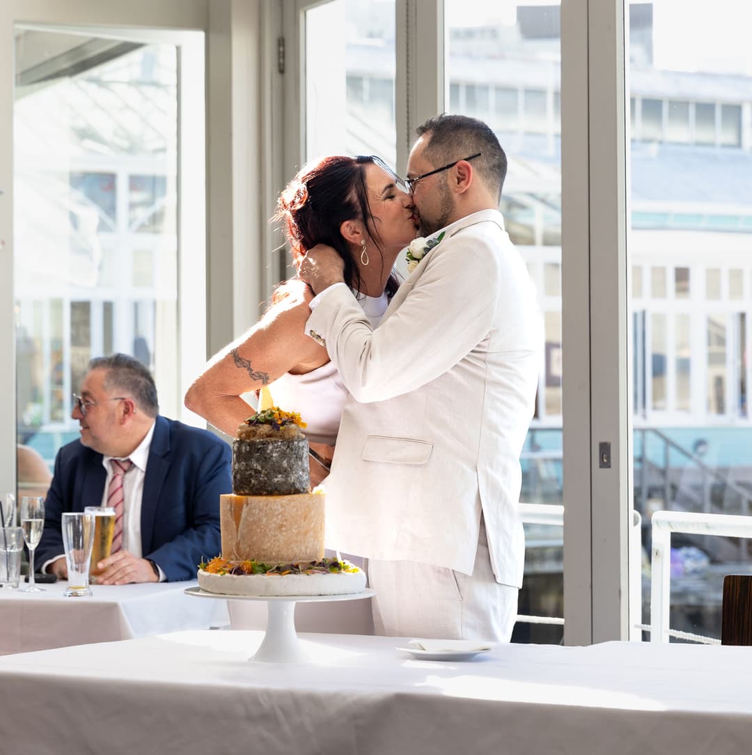 A bride and groom share a kiss in front of a window at their wedding reception, with a two-tier cake on the table nearby and a guest seated in the background.