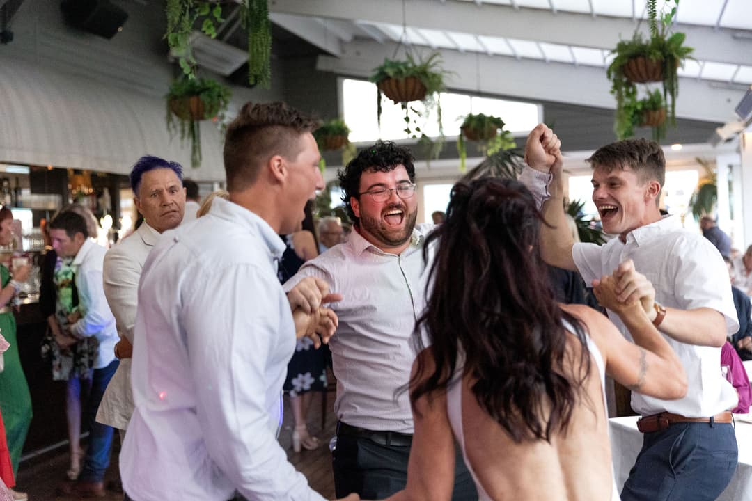 A group of people joyfully dance in a circle at an indoor event, smiling and holding hands. Green hanging plants decorate the venue, and other guests are visible in the background.