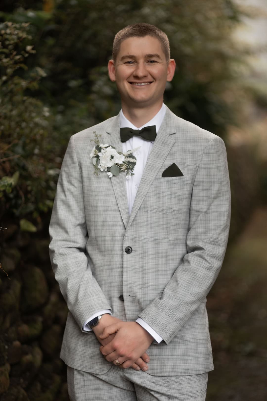 A young man stands outdoors wearing a light grey suit, black bow tie, and a white buttonhole, smiling with his hands clasped in front of him. Greenery and a stone wall are blurred in the background.
