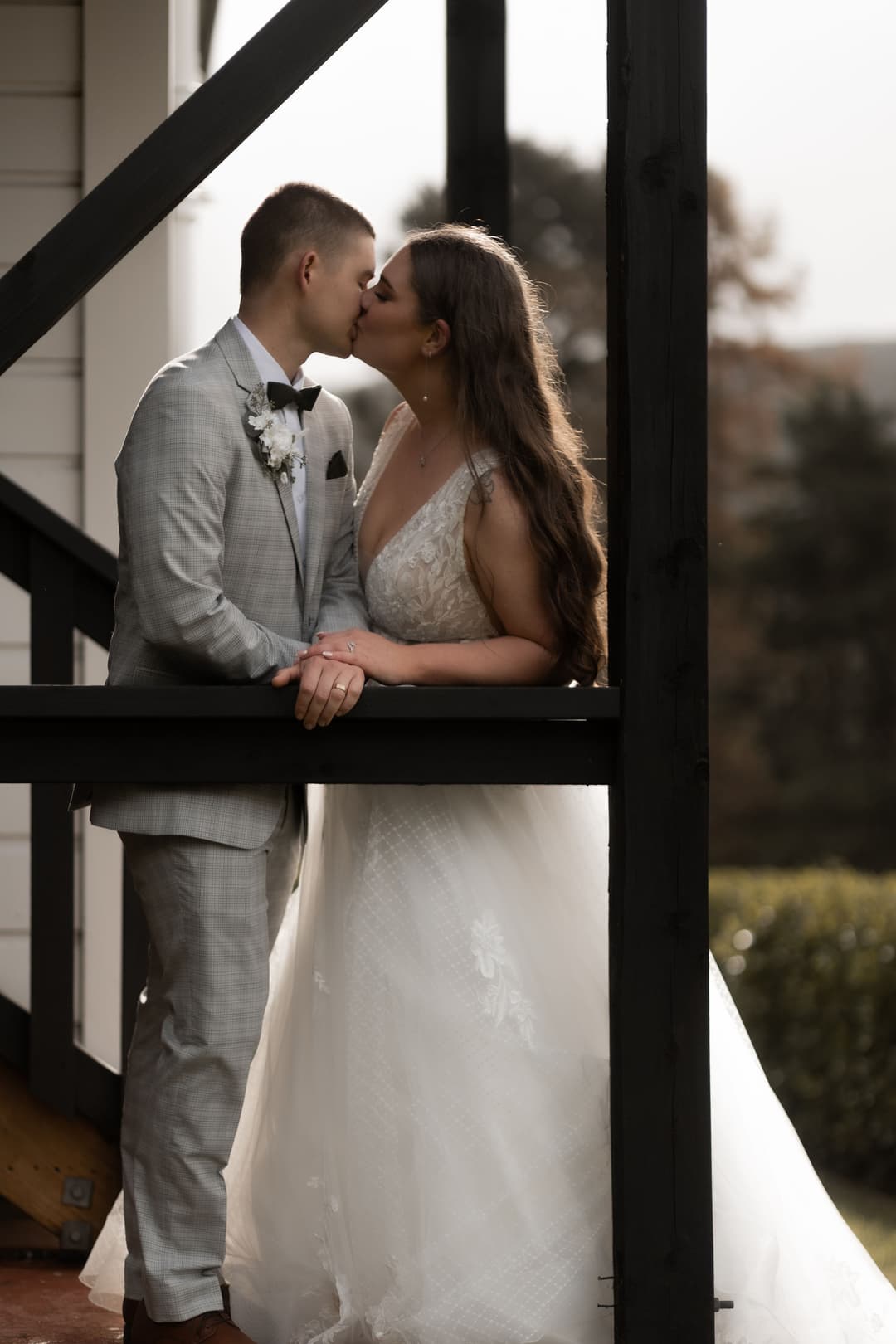A bride in a white gown and a groom in a light grey suit kiss while standing on an outdoor wooden veranda, surrounded by soft natural light.