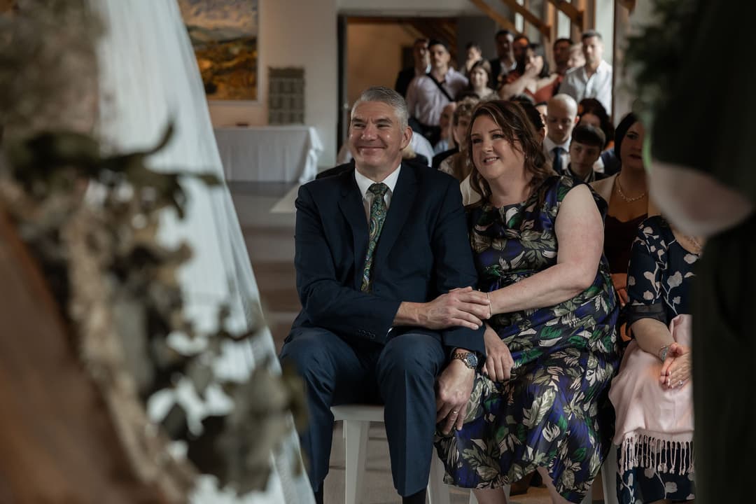 A smiling couple sit closely together in the front row at a wedding ceremony, holding hands, with other well-dressed guests seated behind them. The atmosphere appears joyful and intimate.