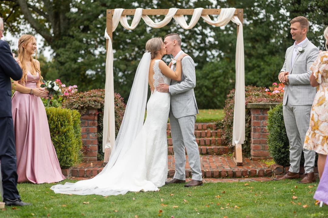 A newlywed couple kisses under a draped wooden archway outdoors, surrounded by greenery and brick steps, whilst smiling wedding guests stand nearby.