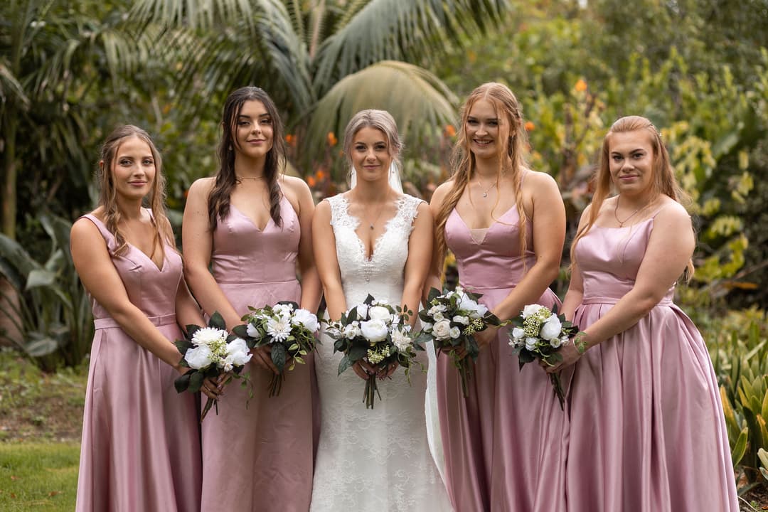 Five women stand together outdoors; the bride is in the centre wearing a white lace dress, and four bridesmaids in matching pink dresses hold white flower bouquets. Lush greenery surrounds them in the background.