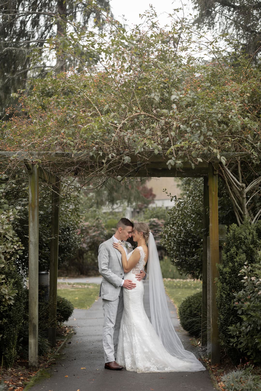 A bride and groom embrace and kiss under a wooden garden arbour surrounded by greenery. The bride wears a white lace gown and long veil; the groom wears a light grey suit. The scene is serene and romantic.
