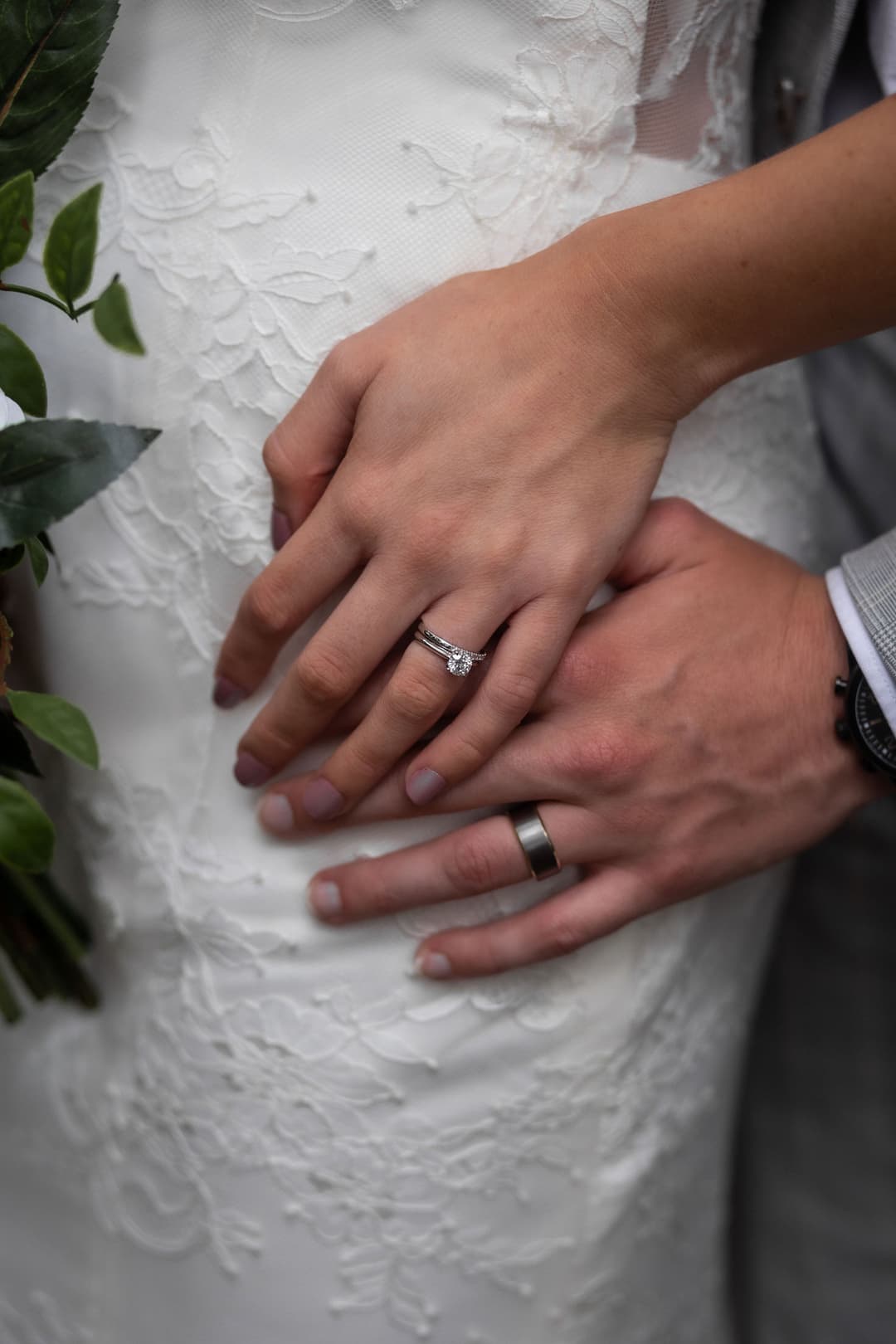 Close-up of two hands resting on a white lace wedding dress; one hand wears an engagement ring and the other a wedding band, symbolising a newlywed couple. Green leaves are partially visible on the left.