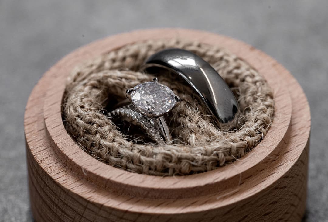 Two rings—a diamond engagement ring and a plain silver wedding band—rest on a coil of jute rope inside a small, round wooden box with a light wood finish.