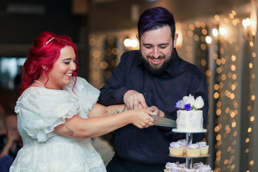 A smiling couple with brightly coloured hair cut a small, decorated cake together at a celebration. The woman wears a white dress, and cupcakes are arranged on a tiered stand beside them. Fairy lights glow in the background.