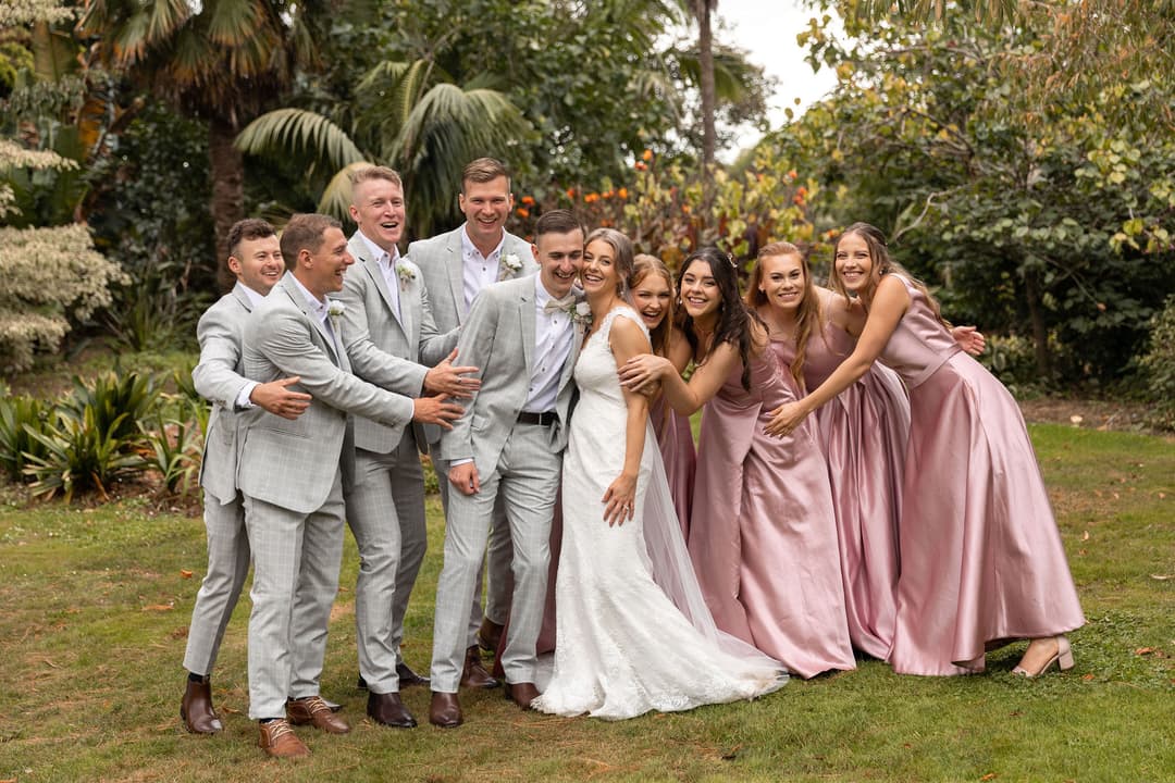 A wedding party poses outdoors, smiling and laughing. Four men in light grey suits and five women in long, light pink dresses surround a bride in a white gown and groom in a grey suit, standing close together on green grass.