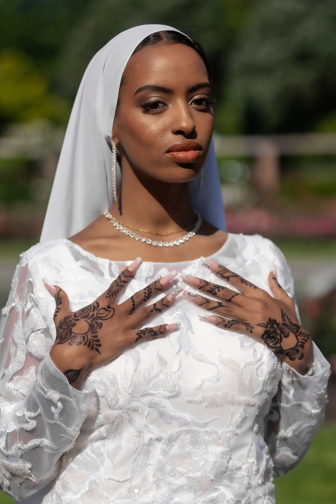 A woman in a white dress and veil stands outdoors, displaying intricate henna designs on her hands. She wears a pearl necklace and earrings, with a confident expression. The background is blurred greenery and sunlight.