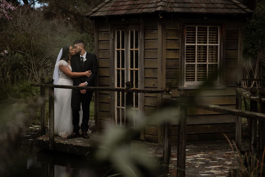 A bride and groom share a kiss while standing on a small wooden bridge beside a rustic wooden cabin surrounded by greenery and water.