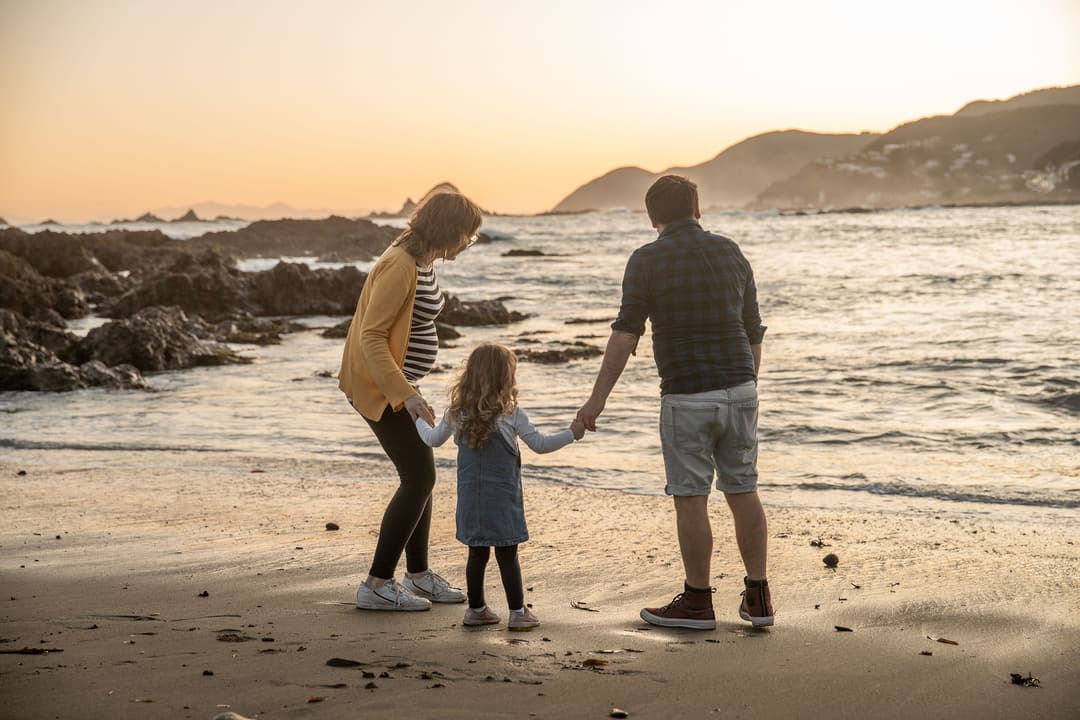 A family of three, two adults and a child, hold hands and stand on a sandy beach at sunset, facing the sea with rocky hills in the background.