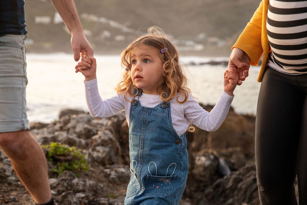 A young girl with long blonde hair in denim dungarees holds hands with two adults by the rocky seaside; one adult has a visible pregnant bump. The group stands outdoors with the sea and hills in the background.