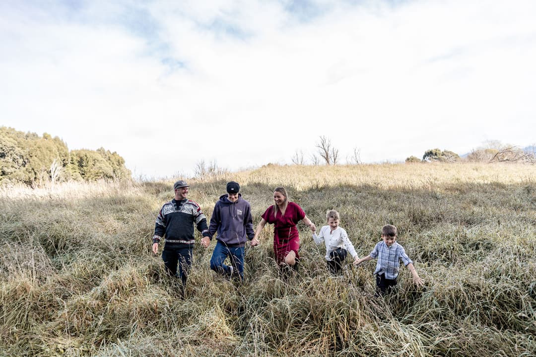 Five people, including two adults and three children, walk hand in hand through a grassy field under a partly cloudy sky, surrounded by trees in the background.
