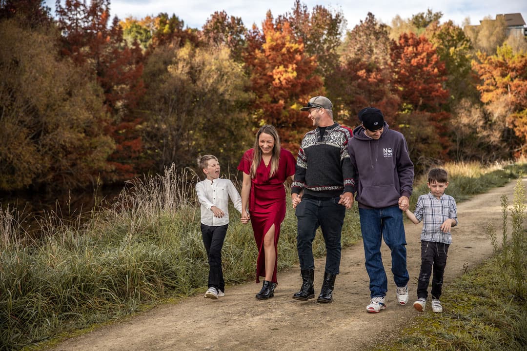 A family of five walks together on a dirt path surrounded by autumn trees. Two adults and three children hold hands and smile, enjoying the outdoors dressed in casual and semi-formal clothes.