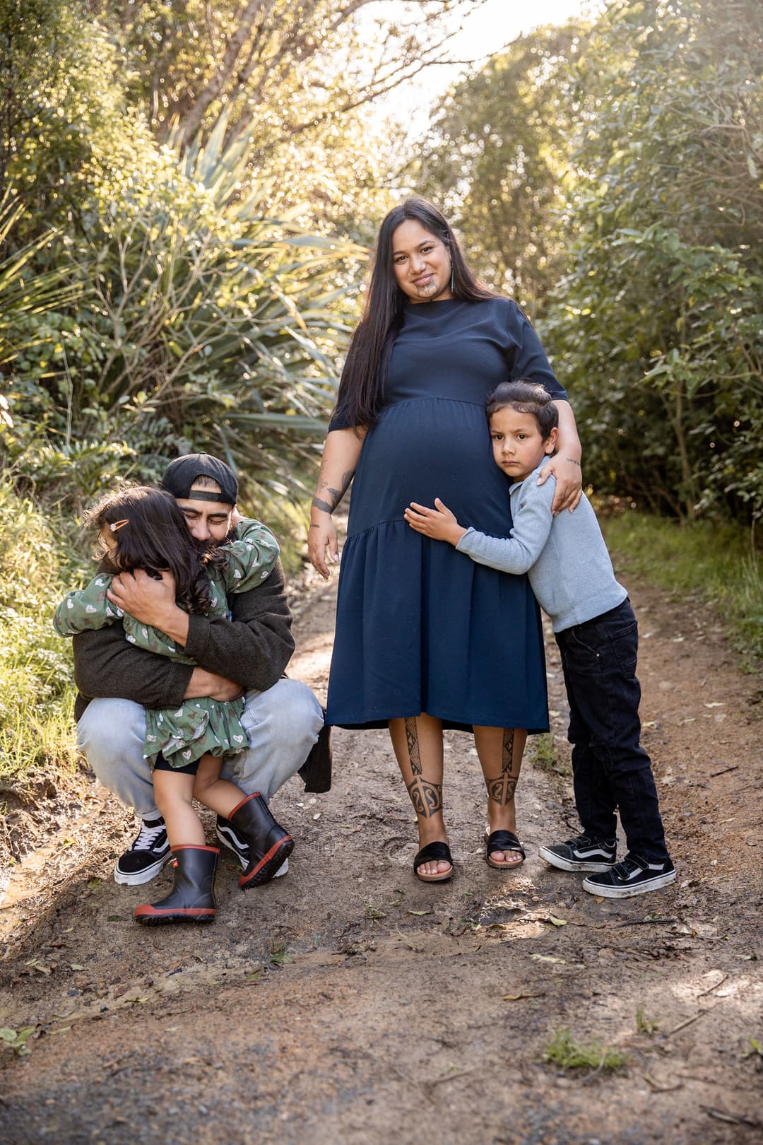 A pregnant woman stands on a path, smiling, with a young boy hugging her. Next to her, a man kneels, hugging a young girl in a green dress. The family is surrounded by greenery and sunlight.