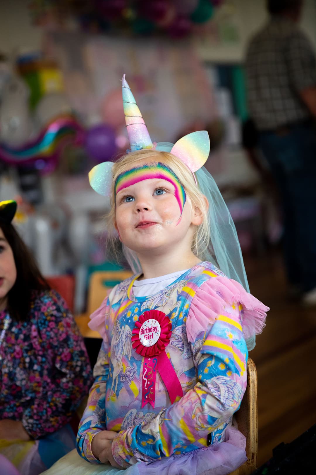 Young girl in unicorn costume with a Birthday Girl! badge, looking up and smiling.