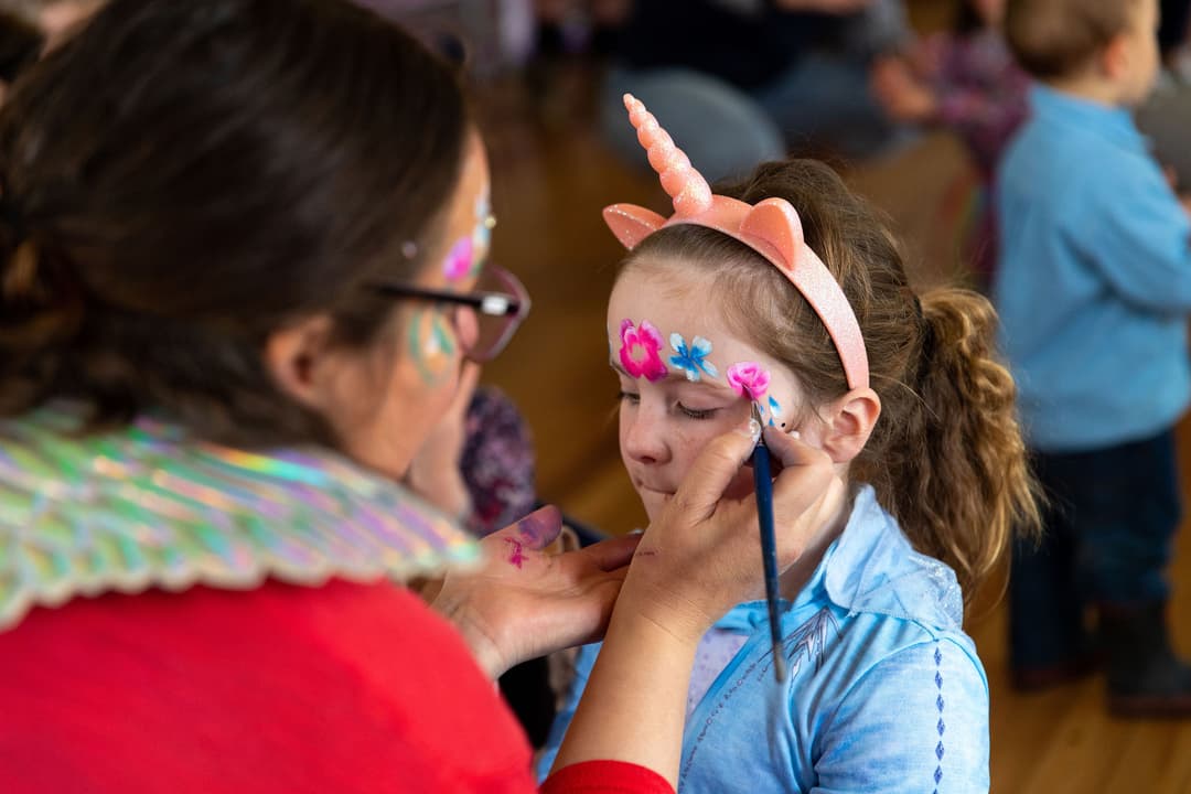 Adult painting a unicorn and flowers on a young girl's face at a party or event.