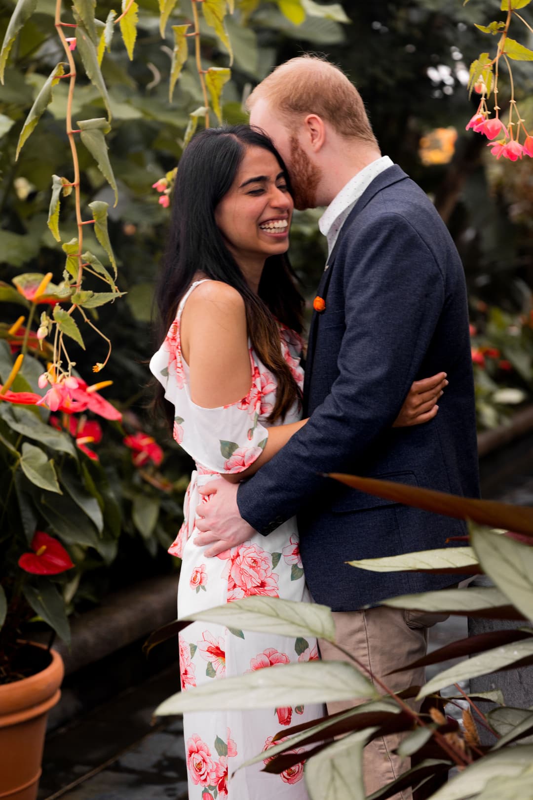A couple embraces and smiles amid lush plants and vibrant flowers in a garden.