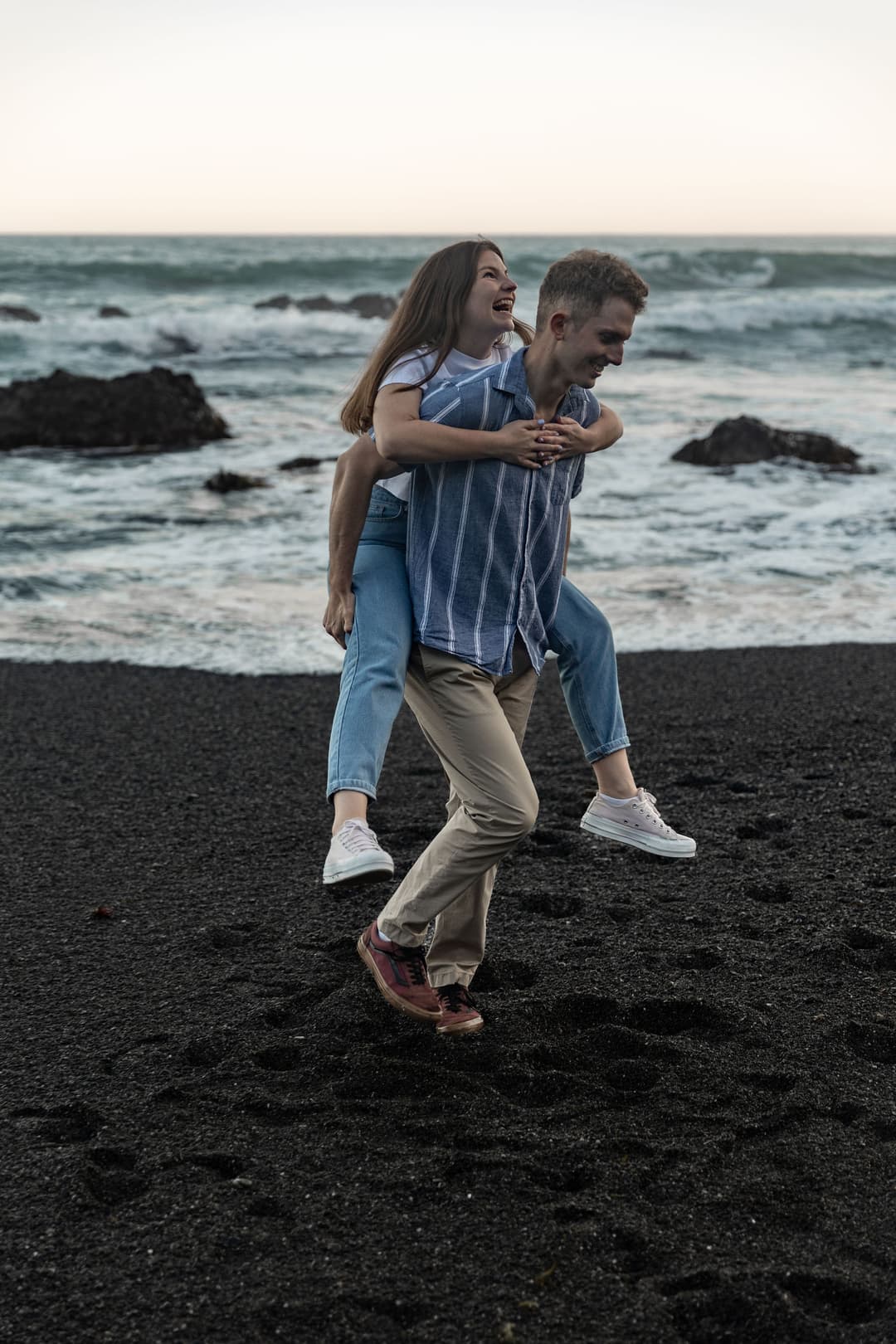 Man giving a woman a piggyback on a rocky beach, both laughing and smiling.