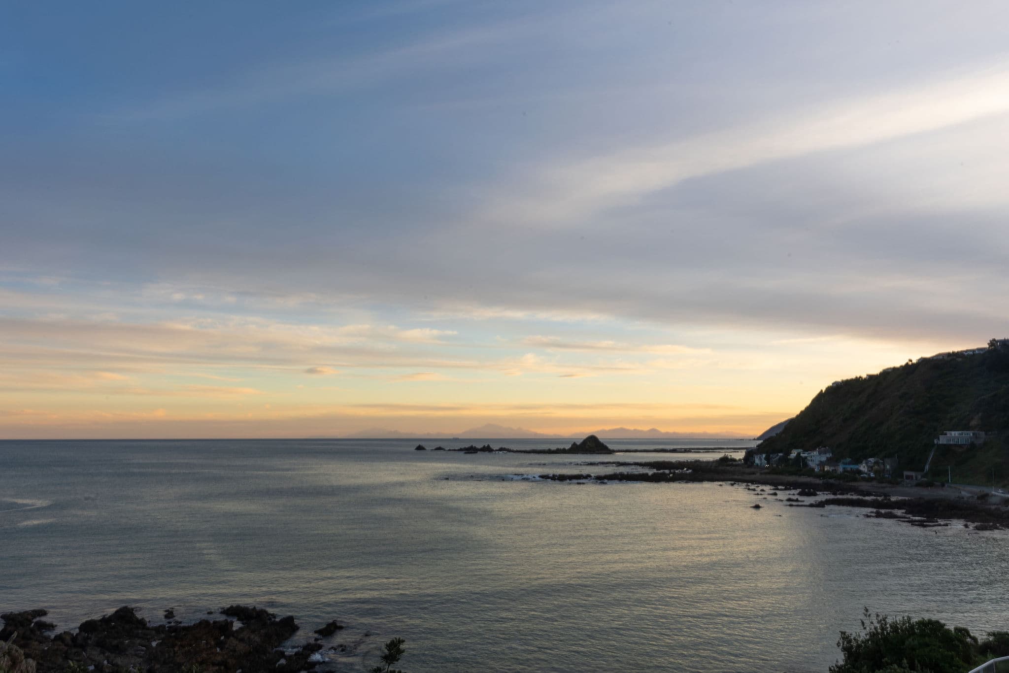 A calm coastal scene at sunset with gentle waves and a hillside on the right.