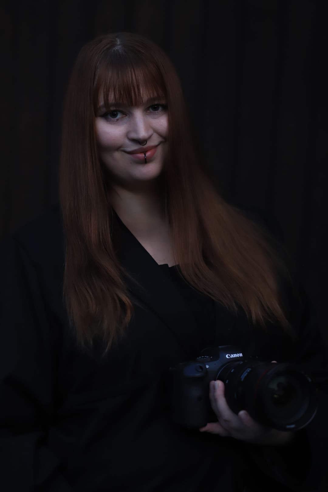 Photographer with long brown hair in a black outfit, holding a Canon camera against a dark background.