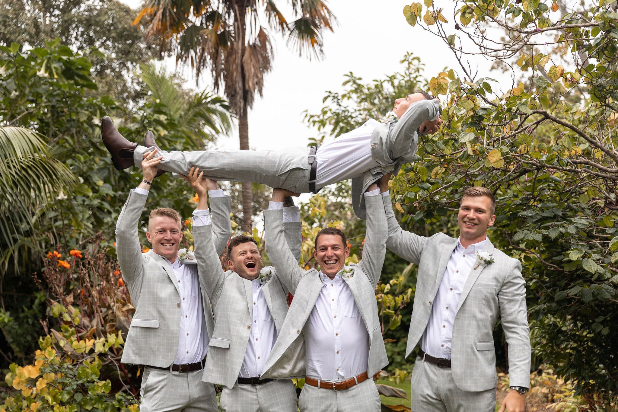 Four men in light grey suits lift another man horizontally in a garden, all smiling and laughing.